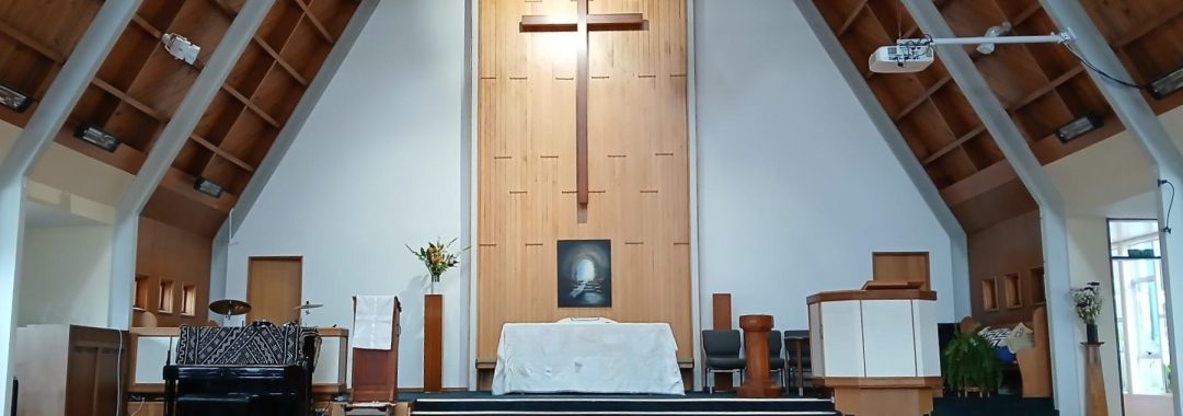Main church auditorium, showing a large A-frame building with a varnished cork floor. There is a large cross above the stage.