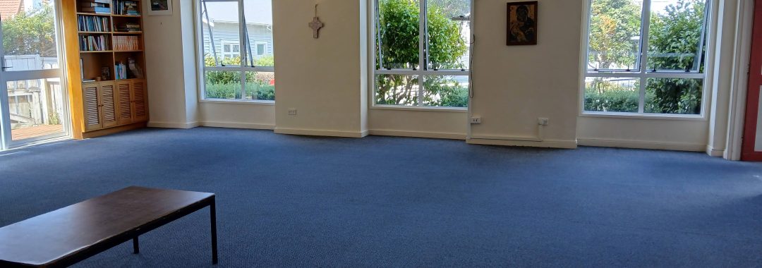 The lounge of the Church complex, showing large wide carpet-covered area and three large windows with triangle tops. There is a small table in the foreground.